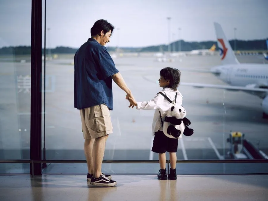 A father and son holding hands, watching planes through the terminal window