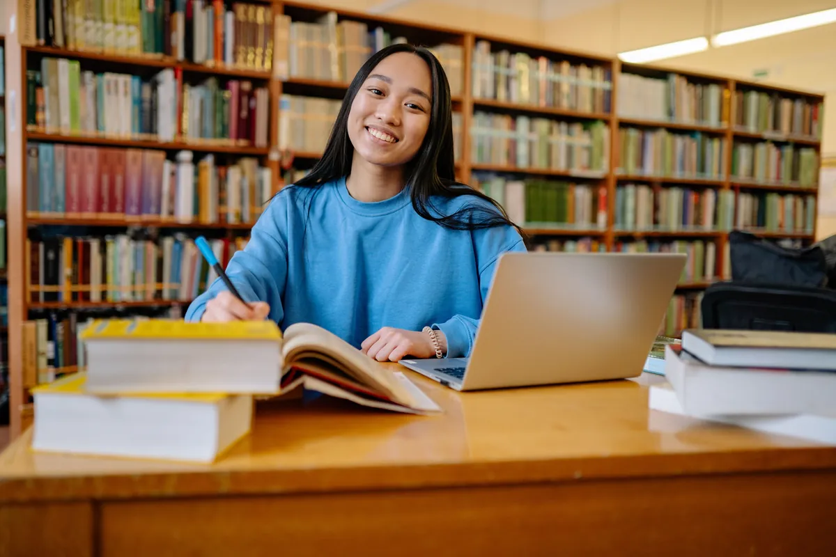 A student smiling at her desk in the library with her laptop and notebooks