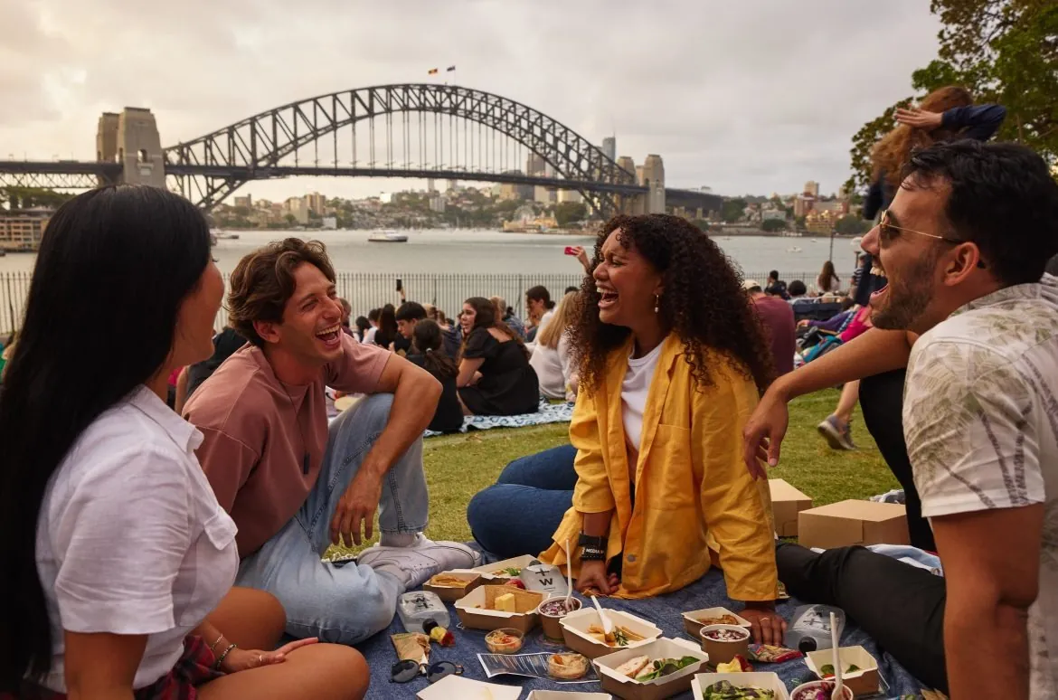 Friends laughing together at a picnic with Sydney Harbour Bridge behind them