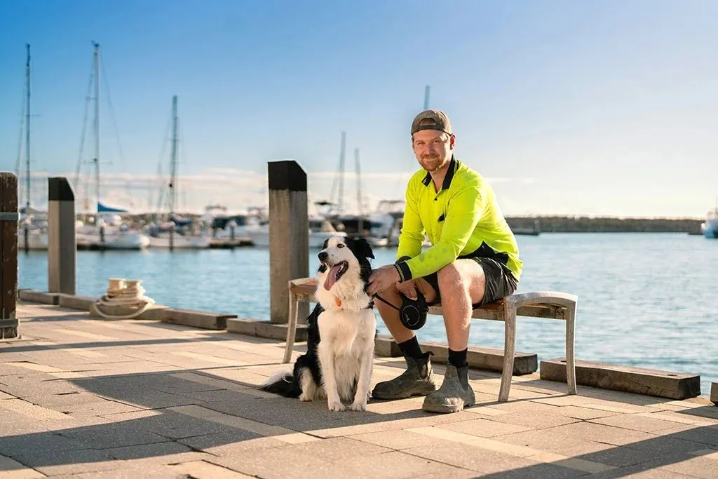 A man in hi-vis sitting on a marina bench with his dog