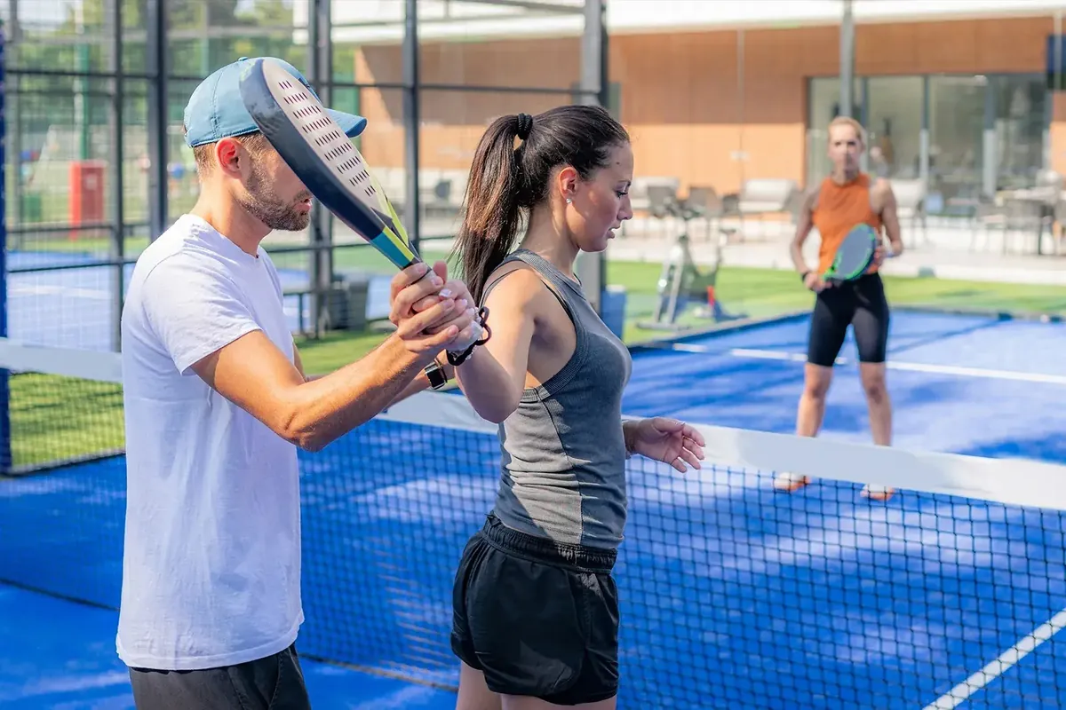 A padel coach guiding a student's racket swing at the net during a lesson