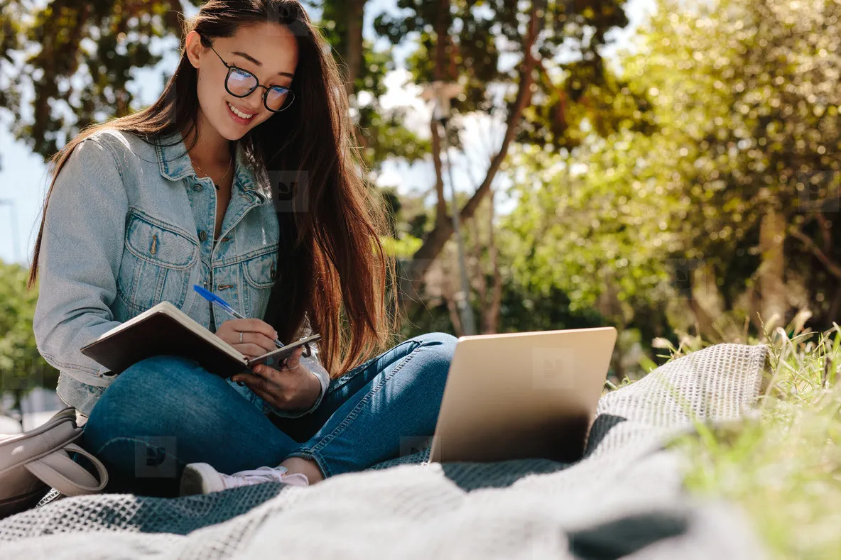 A student smiling over her notebook and laptop in a sunlit park